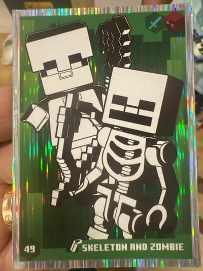The image shows a collectible card featuring two characters from the video game Minecraft. The characters are a skeleton and a zombie, depicted in black and white pixelated art against a green, textured background that resembles grass blocks. The skeleton is on the left, with its ribcage and skull clearly visible, holding what appears to be a bone weapon. The zombie is on the right, also in a pixelated style, with its distinct blocky features. The card has a holographic border that reflects light. In the top right corner, there are small icons representing a sword and a red block, possibly indicating game elements. At the bottom left, the number "49" is displayed, and to the right of that, the text "SKELETON AND ZOMBIE" is written. The card is being held by a hand, with part of a ring visible on the ring finger. The background behind the card is blurred and appears to be an indoor setting. No location in Mainburg, Germany is depicted or suggested by the image.