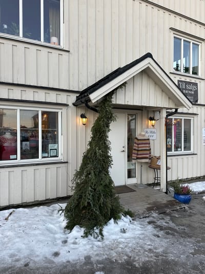 The image displays the exterior of a two-story building in Henningsvær, Norway, during winter or early spring conditions. The building is constructed with off-white, vertical wooden siding.
The ground floor features a central entrance under a dark-roofed gable awning, flanked by large display windows. Two illuminated, black-cased wall lanterns are mounted on either side of the white entrance door. Leaning against the wall, partially beneath the awning, is a large, conical evergreen tree, its base situated in a patch of dirty snow. To the right of the door, a mannequin torso displays a multicolored, patterned knitted sweater. Adjacent to the mannequin, a small round table holds a woven basket and a white sign with black text, partially visible as "Ho Milla." Further to the right, a blue ceramic pot contains small green plants and pink/purple flowering heather.
The ground in front of the building is covered in patches of dirty snow and slush, with some areas of exposed asphalt or concrete visible. Faint tracks are discernible in the snow.
On the upper right side of the building, a rectangular black sign with white text reads "Til salgs" (For Sale) and displays a phone number "75 57 74 00," with the text "AVANTI FISKERS" partially visible below. The building has multiple windows on both floors, reflecting elements of the surrounding environment. No individuals are visible in the scene.