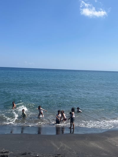 In Batulayar Barat, Indonesia, on a sunny day with a clear blue sky and a few wispy clouds, a group of approximately seven children are playing in the shallow ocean water. The water is a deep blue, with gentle waves lapping at the dark, sandy shore. The children, appearing to be of various ages, are scattered across the frame, some wading, others splashing, and at least one girl appears to be moving towards the shore. One boy in the foreground on the left is wearing patterned shorts and is splashing in the water. To his right, two girls are standing close together in the water, one with her arm outstretched. Further to the right, a girl in a white top and shorts stands on the shore, looking out at the water. In the background, a few other children are visible in the water, some partially submerged. The scene captures a moment of youthful recreation on a tropical coastline.