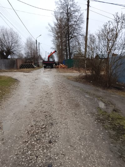 The image depicts a rural road in Высокиничи, Russia, likely after rainfall, judging by the wet, gravel-covered road surface and scattered puddles. In the middle distance, a dark-colored truck with an orange crane arm is parked on the side of the road. Two people are visible near the truck, possibly involved in loading or unloading items. To the left of the truck are stacked concrete pipes, and to the right, there are piles of cut wood. Fences and trees line both sides of the road, suggesting a residential area. Overhead utility lines are visible against an overcast sky.