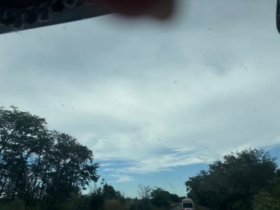 The image depicts a view from inside a vehicle, looking out at a cloudy sky over a landscape of trees and a road. On the left side of the image, a dense cluster of dark green trees is visible. In the distance, a light-colored vehicle, possibly a van or camper, is seen on the right side of the road. The road itself is paved and appears to be in a rural or suburban area, with vegetation lining its sides. The sky is predominantly filled with thick, gray clouds, though patches of blue sky with lighter clouds can be seen, particularly towards the horizon. The overall impression is that of a somewhat overcast day, possibly in the late afternoon or early morning, given the lighting. The scene is likely captured while driving, as suggested by the perspective and the blurry details of the sky and surroundings. The image has a number of small dark spots, possibly dust or water droplets on the windshield. There is no discernible city infrastructure visible. The image may have been taken in the vicinity of Cherni Vrah, Bulgaria, but this cannot be confirmed from the visual evidence alone.