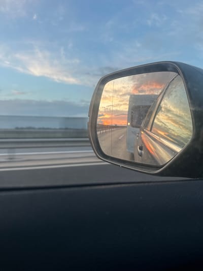 The horizontally oriented image captures a detailed view of a car's side mirror reflecting a vibrant sunset over a road. The mirror, which shows signs of dust or streaks on its surface, offers a clear reflection of the sky ablaze with warm orange, yellow, and red hues as the sun sets low on the horizon. The reflection also shows a long stretch of road extending into the distance, with faint glints of light, possibly from oncoming traffic or distant structures.
Outside the mirror, to the left, a clear blue sky with sparse, light clouds is visible, suggesting a calm weather day. To the right of the mirror, a dark, textured portion of the car's interior, likely a door panel, frames the right edge of the composition.
The overall scene depicts a moment during late afternoon or early evening, characterized by the intense colors of the sunset. The perspective is from inside a vehicle, either stationary or in motion, with the reflection providing a captivating view of the landscape behind and to the side. There is no discernible text visible in the image. The location context for this scene is Óföldeák, Hungary.