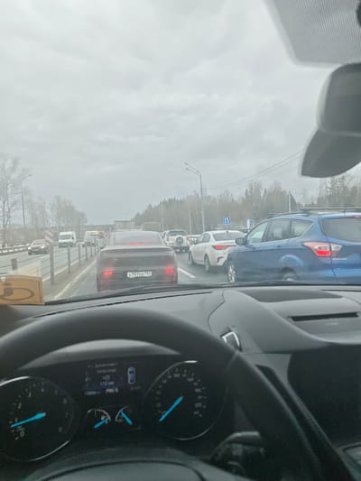 The image provides a first-person view from inside a car in a traffic jam on an overcast day, likely in Sukharevo, Russia.

In the foreground, the top of a black steering wheel is visible, along with parts of the dashboard. The instrument cluster shows a speedometer and tachometer with blue needles. A digital display indicates "112 км" (kilometers), "118984 км" (odometer reading), and "P R N S" for gear selection. A yellow object with an indistinguishable symbol rests on the dashboard near the windshield.

Through the windshield, a multi-lane highway or major road is seen filled with numerous cars. The vehicles are closely packed, indicating slow-moving or stopped traffic. Directly ahead in the same lane is a dark gray sedan with a Russian license plate "X797EE797". To the right, a blue Ford Escape SUV is visible, its rear "ESCAPE" badge identifiable. Other vehicles include white sedans, a white van, and various SUVs and trucks.

The sky is uniformly gray and overcast, suggesting a cloudy day without direct sunlight. Bare trees line the roadside in the background, implying late autumn, winter, or early spring. Road infrastructure such as lampposts and road signs with Cyrillic text are also present along the highway. The overall scene depicts a busy road in an urban or suburban setting during daytime.