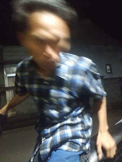 The image captures a person, likely male, in a dimly lit environment, possibly at night or indoors. The individual is wearing a blue and white plaid button-up shirt and blue pants. Their face and upper body are significantly blurred due to motion, making specific features indiscernible. Their right arm is visible, with the hand resting on a dark object, which could be a vehicle seat or part of a motorcycle, suggesting the person might be sitting on or leaning against it.
The background is dark and indistinct, showing faint architectural elements such as walls on the left and right. On the right-hand wall, a small, dark-framed item, possibly a picture or mirror, is visible. The floor appears dark and concrete-like. The overall lighting suggests a low-light condition, contributing to the blurry effect. No discernible emotions or visible text are present. The scene is located in Kecamatan Jangkar, Indonesia.