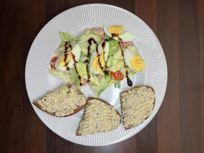 A top-down, medium shot captures a white, fluted plate holding a salad and three open-faced sandwiches on dark bread. The salad, in the upper portion of the plate, consists of chopped lettuce, sliced tomatoes, and hard-boiled eggs, all drizzled with a dark sauce. The three sandwiches, arranged in the lower portion of the plate, are topped with a creamy spread containing visible pieces of tuna and red onion. The plate rests on a dark, wooden surface. The image is undated and lacks any identifiable location in Balbriggan, Ireland.