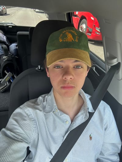 The image shows a young man wearing a blue and white striped shirt with a small polo logo on the left breast. He is wearing a green and brown cap with an embroidered logo that reads "British Falconers Club" and an owl design. He is sitting in the passenger seat of a car with a black seat belt fastened across his chest. The car is parked on the side of a road, with another vehicle visible in the background. The man is looking directly at the camera with a neutral expression on his face. The image appears to be taken in a vehicle in the Kirklees area of the UK.