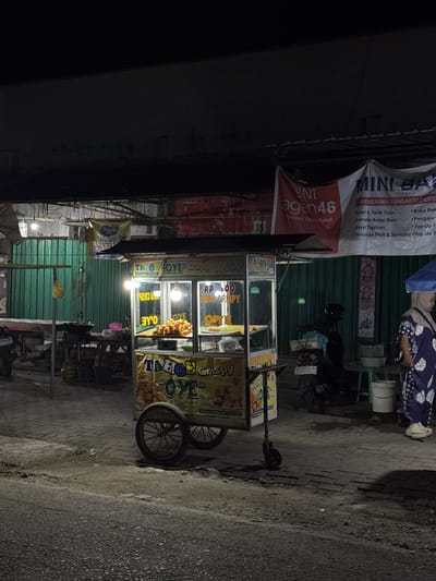 The image depicts a street scene at night in Alian, Indonesia. A food cart selling "Tahu Oye Crispy" is prominently featured. The cart has a glass display showcasing fried tofu and signage with prices and product names. A woman in a patterned dress stands to the right of the cart. Various other elements of the street environment are visible, including a motorcycle parked behind the cart, tables, green corrugated metal walls, and streetlights. Banners advertising "BNI Agen46" and "MINI BANK" are hung above the food cart. The ground is paved with bricks and asphalt.