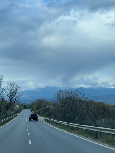 The image depicts a two-lane asphalt road leading through a rural area near Ribnik, Bulgaria, under an overcast sky. A black SUV is observed traveling away from the viewer in the right lane of the roadway. The road is marked with a continuous white line on the right shoulder and dashed white lane dividers. Metallic guardrails, fitted with red reflectors, run along both sides of the road. The immediate environment consists of dense deciduous vegetation, including trees and bushes, many of which appear bare or in early foliage. In the background, a range of mountains with visible snow patches on some peaks is partially obscured by low-lying clouds. The sky is largely covered by heavy, grey cloud formations, with minor patches of lighter blue visible at the top. No people are visible.