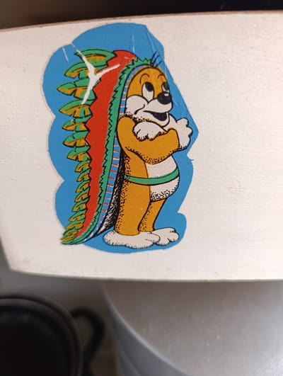The image shows a close-up of a sticker on a white surface, likely painted wood. The sticker features a cartoon character that appears to be a porcupine with its quills stylized to resemble a headdress or mane. The character is orange with white accents, black eyes, and a black nose. It has its arms crossed. The quills are multi-colored, with layers of green, orange, and red, outlined in black and green. A green band encircles the character's waist. The background of the sticker is a bright blue, cut into a scalloped shape. The sticker has some damage, with a noticeable tear and white areas showing where the sticker has peeled or is missing. The white surface beneath the sticker has a textured appearance. The lighting suggests an indoor setting, with no indication of time of day or weather. There is no visible text in the image. The lower portion of the image shows a blurry view of a black object and a white curved surface, possibly part of the object the sticker is attached to.