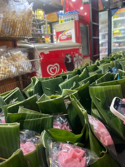 In the city of Barat Yaman, Indonesia, an image captures a close-up view of numerous food items individually wrapped in banana leaves, arranged in rows. Each package is secured with a toothpick. A few of the packages reveal glimpses of pink, textured food items, possibly snacks or sweets, encased in clear plastic. In the background, the scene transitions to a store setting. A red refrigerator with an "Algida" logo and a heart symbol is visible, along with shelves stocked with various products, including bottled water. Another refrigerator to the right is also filled with items. The overall ambiance suggests a bustling market or a small shop.