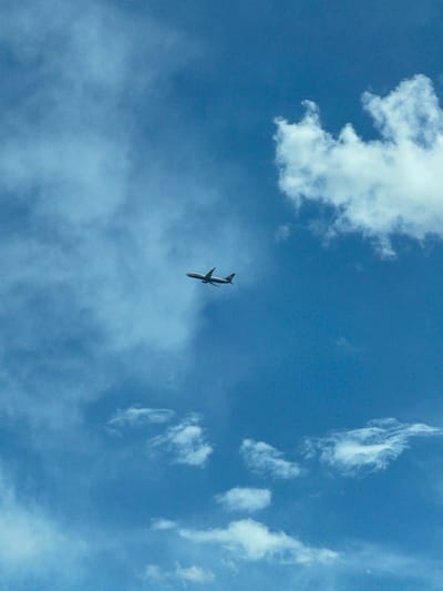 The image shows an airplane flying in a blue sky with scattered clouds. The plane is centrally located in the frame, viewed from below, with its belly facing the viewer. It's a daytime shot with natural lighting. No people are visible. The location of the photograph in Laka, Bulgaria is not discernible from this image.