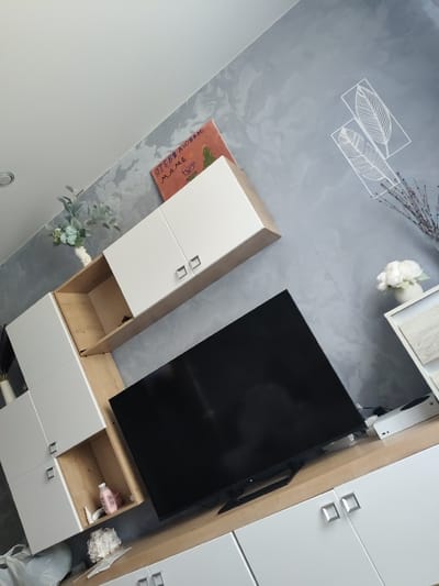 The image shows a modern living room with a wall-mounted entertainment unit and a television. The unit consists of white cabinets with wooden accents and open shelving. A black flat-screen television is placed on the lower wooden surface. Decorative items like a vase with artificial flowers, a framed artwork with text in Russian that translates to "I love you, Mom" and a Christmas tree, and two framed feather outlines are visible on the wall and shelves. The wall has a textured gray finish. The overall scene depicts a well-organized and contemporary living space. No specific details about Shimanovsk, Russia are present in the image itself.