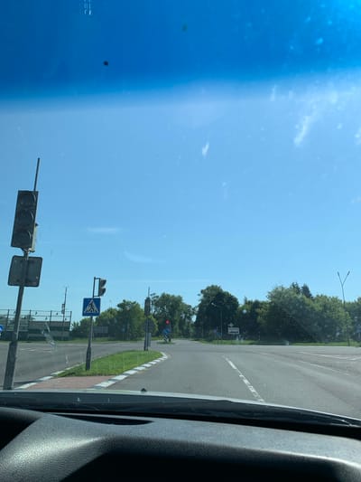 The image shows a view from inside a vehicle looking out through the windshield at a road intersection on a clear, sunny day. The road is paved and marked with white lines. There are traffic lights, pedestrian crossing signs, and trees visible. A sign in the distance appears to be a town or village name, "Шерешевцы". The foreground shows the dashboard of the vehicle.