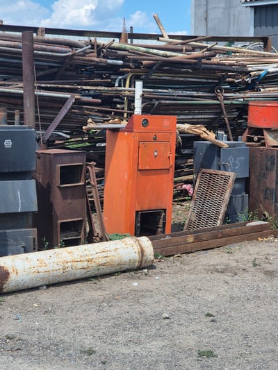 The image depicts an outdoor scene at what appears to be a scrapyard or industrial storage area in Pidhaitsi, Ukraine. In the foreground, a large, white, rusty pipe lies horizontally on the ground. Behind it, a collection of various metal objects are visible, including dark grey rectangular containers stacked on the left, a rusty, box-like structure with openings, and an orange industrial cabinet or boiler with a dial and a door. To the right of the orange cabinet, a weathered metal grate leans against a dark rectangular object. In the background, a massive pile of assorted metal pipes, some rusty and others painted in various colors including green and blue, are stacked high against a building structure. The ground is covered in gravel and dirt, with sparse green vegetation growing in places. The sky above is partly cloudy with a blue sky.
