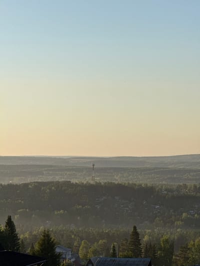The image, when correctly oriented, presents a vast panoramic landscape, likely captured from an elevated viewpoint. The upper half of the scene is dominated by a clear sky, transitioning smoothly from a light blue hue in the upper left to a softer, yellowish-orange tone near the horizon, suggesting either sunrise or sunset.
A distinct, undulating horizon line stretches across the mid-section of the image, separating the sky from the expansive land below. Along this distant horizon, a tall communication or cell tower stands prominently, rising slightly above the tree line.
The lower half of the image is covered by a dense, verdant forest, stretching into the distance. The foliage appears dark green with lighter, brighter patches, indicative of sunlight filtering through the trees. Scattered within this forested area, particularly in the middle ground, are numerous small buildings or houses, partially obscured by the trees and a soft atmospheric haze, suggesting a village or scattered settlement integrated within the natural environment.
In the bottom right corner of the foreground, parts of a dark-colored roof and some closer, larger trees are visible, indicating the vantage point is from or near a building.
The scene conveys a sense of tranquility and vastness. The soft lighting and slight haze over the forest suggest either an early morning or late afternoon setting. There is no discernible activity taking place, and no visible text is present in the image. The location is Minino Stantsiya, Russia.