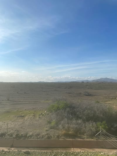This wide-angle landscape photograph, taken in Jbilate الجبيلات, Morocco, presents a vast natural scene under a bright, clear sky. The image is vertically oriented, with the sky occupying the left side and the land the right.
The left half of the frame features an expansive, vibrant blue sky, indicative of fair weather. Scattered across this azure expanse are delicate, wispy white cirrus clouds, appearing as thin, elongated streaks that add subtle texture without obscuring the clear view.
The right half of the image depicts a dry, somewhat arid landscape. In the immediate foreground, along the bottom edge, there's a distinct concrete or earth embankment, possibly a roadside feature, bordered by sparse, light green grass. A metal structure, resembling a fence post with several wires, is also visible on the far right. Moving further into the scene, the midground is characterized by dark, dry earth, with visible patterns that could be erosion or traces of past agricultural activity. This area is dotted with patches of low-lying vegetation, ranging from scattered light green grasses to denser, darker scrub bushes. In the background, a series of hazy, undulating hills or mountains stretches across the horizon, their forms softened by distance and atmospheric perspective.
No people or explicit activities are depicted, giving the scene a serene and untouched quality. The strong, clear lighting suggests it is daytime, likely morning or early afternoon. The arid terrain, sparse vegetation, and distant mountains are consistent with the geomorphology of the Jbilate region. There is no visible text within the image.