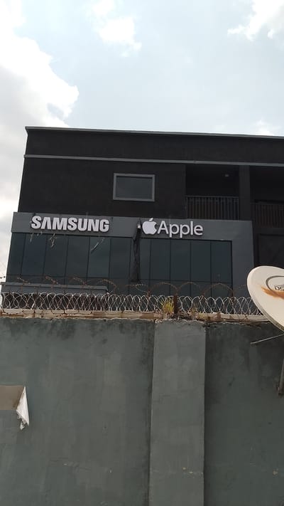 The image shows the exterior of a two-story building in Effurun, Nigeria, with the signage "SAMSUNG" and "Apple" displayed prominently above a row of windows. A gray concrete wall topped with barbed wire runs in front of the building. Part of a satellite dish is visible on the right side. The building is painted in a dark gray color. The sky is overcast with clouds.