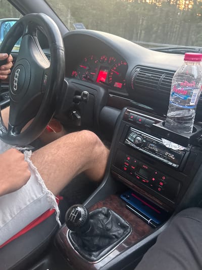 The image captures a partial view of a car's interior, likely an Audi due to the visible logo on the steering wheel and an "S line" badge. The perspective is from the passenger side, looking towards the driver and the dashboard. The driver's hand is on the steering wheel, and their bare legs, wearing white frayed shorts with a red lining, are visible.
The dashboard is illuminated with red lights, displaying analog gauges for speed, RPM, fuel level, and engine temperature. A digital readout shows an odometer reading of 248,832 km, a trip meter of 519.2, and an outside temperature of 19.5°C. An aftermarket Alpine brand car stereo is installed in the center console, displaying "87.50" MHz (likely an FM radio frequency) and "F16". Below it, the climate control panel features buttons such as "AUTO", "TEMP", "ECON", and "OFF".
A bottle of "SKA Spring water" with the website "www.lina07.com" printed on its label is placed in a cup holder. The car features dark wooden trim on the center console. A manual gear shifter with a carbon fiber-look knob clearly shows the 5-speed and reverse (1, 2, 3, 4, 5, R) pattern. Tucked next to the shifter are a smartphone in a blue case and what appears to be a vape pen. Golden sunlight streams through the windshield, indicating either early morning or late afternoon, suggesting the car is outdoors in a sunny environment, consistent with the provided location context of Dzhebel, Bulgaria. The high mileage suggests a well-used vehicle, likely an older model Audi.
