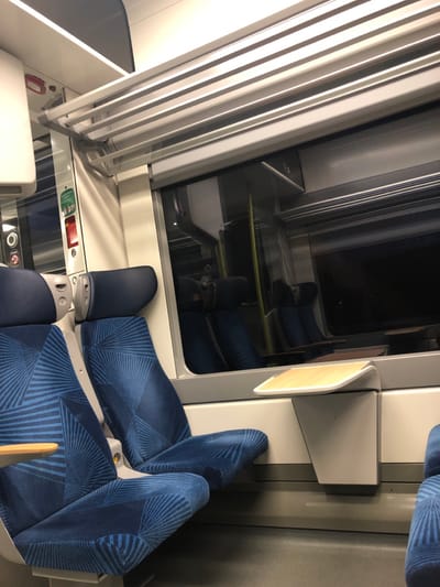 An interior shot of a train car shows blue seats with a geometric pattern. A window with partially closed blinds reflects additional seating. Each seat has a small, fold-down table attached to the side wall. The composition is rotated counter-clockwise, creating a slightly angled perspective of the train's interior.
