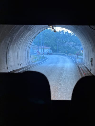The image shows the view from inside a concrete tunnel, looking out towards the exit. The tunnel has a rounded arch ceiling with lights mounted along its side. The road inside the tunnel is paved and appears to be wet, with small white particles scattered on its surface.
Beyond the tunnel exit, a paved road curves to the right. A tanker truck is visible driving on this road. The surrounding environment consists of a hillside covered in trees and vegetation, with some buildings and a power line tower in the distance. Several road signs are present, including a warning sign for a curve to the right and a speed limit sign.
The foreground of the image is dominated by dark, silhouetted shapes, possibly the interior of a vehicle such as headrests. The overall lighting suggests it is daytime, but potentially overcast or dusk. This scene is located within the vicinity of Gülnar, Turkey, as indicated by the context of the request.