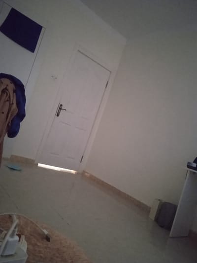The image is of an interior room with white walls and a white door. There is a closed door on the left wall. The camera appears to be lying on the floor looking towards the door. There are no visible people in the image. A light blue object, possibly a rug, is visible on the floor in the foreground. A white table with a white surface on top is in the right corner of the room. A dark object is lying on the table. A gray box is on the floor by the table. The room is located in Réghaïa, Algeria, at the coordinates 36.729 latitude and 3.369 longitude.