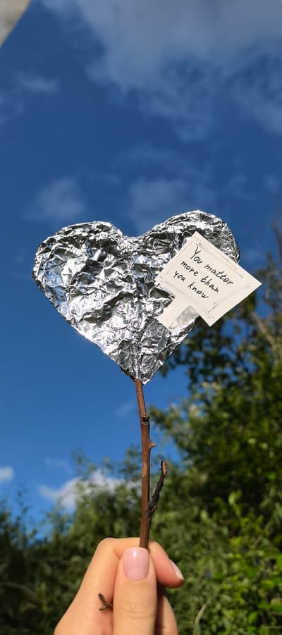 A close-up, vertical shot captures a hand holding a small, twig-like stick. Attached to the stick is a heart-shaped object made of crumpled aluminum foil. A small, square piece of paper, with the handwritten message "You matter more than you know", is affixed to the foil heart. The background is a bright blue sky with scattered white clouds and the blurred green foliage of trees. The scene is outdoors, likely during the daytime, given the clear sky and sunlight reflecting off the foil. No specific location in Villeneuve-le-Roi, France is identifiable from the visual information.