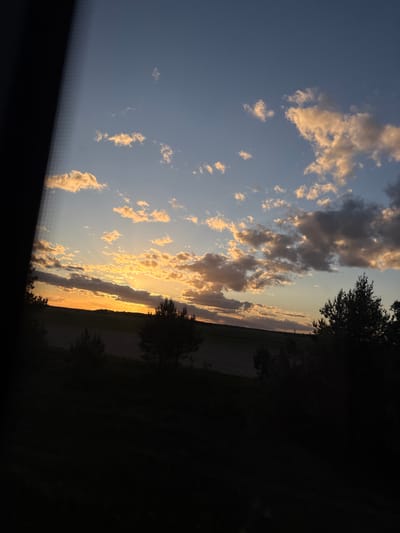 The image shows a sunset in Мажэйкаўскі сельскі Савет, Belarus. The sky is a gradient of blue to orange with scattered cumulus clouds catching the golden light of the setting sun. Silhouettes of trees and vegetation are visible along the horizon line. The foreground is mostly dark.