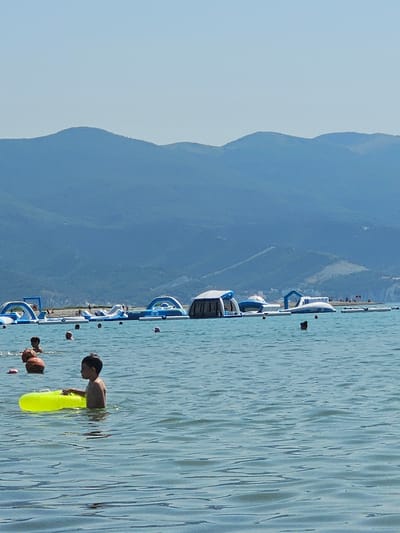 This is a vertical, eye-level, medium shot of a bright, sunny day at an inflatable water park on the water. In the foreground, a young boy with dark hair is partially submerged in the clear blue water. He is facing to the left, looking down at the water, and holding a bright yellow inflatable tube in front of him. His body is angled slightly away from the viewer. To the left of him, another person's head is visible above the water, with a reddish-brown inflatable ring. Further back, a line of colorful inflatable water structures, predominantly white and blue, are arranged on the water. These structures appear to be a water obstacle course or playground. Beyond the water park, a strip of land with some buildings and trees is visible, suggesting a shoreline. In the background, large, imposing mountains with gentle slopes dominate the horizon. The sky is a clear, pale blue. The water is calm with gentle ripples reflecting the sunlight. The overall scene conveys a sense of fun and recreation on a warm summer day in Myskhako, Russia.