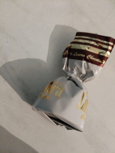 The image shows a single piece of candy wrapped in silver and gold foil, resting on a light-colored, textured surface. The candy is twisted at both ends, with one end featuring a dark red and gold striped wrapper. Text is visible on the striped wrapper, which appears to read "Cafe Aurora Chocolate." The other end of the candy has a silver wrapper with gold lettering that is partially obscured. A shadow is cast to the left of the candy, suggesting a light source from the right. The background is uniformly lit and shows no discernible features of the city of Khmchik, Russia, or any other urban environment. The scene appears to be an indoor setting with minimal background detail.