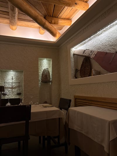 A low-angle, medium shot depicts the interior of a restaurant or similar establishment. On the left, a wooden wall with logs. Above and to the right is an opening in the ceiling with a basket and old, red door. The rest of the walls are textured and cream-colored. Within the walls are two illuminated alcoves, one containing a basket and the other displaying old-fashioned scales. To the right, tables are set with white tablecloths, glassware, and dark chairs. The restaurant is located in Agropoli, Italy.