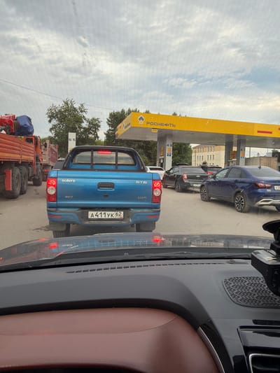 This image depicts a queue of vehicles at a "Rosneft" gas station in Skopin, Russia. The primary focus is on a blue Sollers ST6 pickup truck with license plate A411YK 62, positioned at the front of a line of cars and trucks. The scene appears to be in daylight, under an overcast sky, suggesting a cloudy day.

Other vehicles visible include a dark blue passenger car in front of the pickup, and several trucks behind it, one of which has a blue tarp covering its cargo. The gas station's distinctive yellow canopy with "Rosneft" branding is prominently displayed on the left side of the image, along with pricing information and pump numbers like "43" and "65". The view is from inside another vehicle, looking forward, with parts of its interior, including a brown leather seat and dashboard elements, visible in the foreground. The overall atmosphere suggests a routine stop at a gas station, possibly during a journey.

Visible text includes "РОСНЕФТЬ", "4,5м", "ST6", "2.0CTI", "sollers", "4WD", "А411 УК 62", "RUS", "A678YK", "x58", and "GLB".