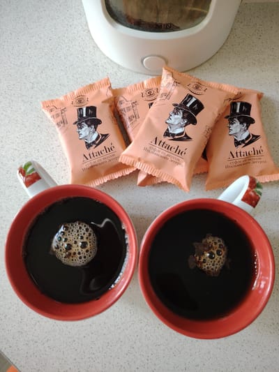 The image shows a top-down view of two red mugs filled with dark liquid, presumably coffee or tea, resting on a speckled countertop. A few peach-colored packets of "Attaché" brand suflé with Italian dessert flavor are scattered on the counter between the mugs. The packets feature an illustration of a man in a top hat. In the background, a portion of a white electric kettle is visible. The scene is illuminated by natural light. There is no indication of the location being in Vladimirovka, Russia.