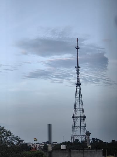 The image shows a tall, lattice-style radio tower in Karnal, India, silhouetted against a cloudy sky. The tower is segmented with platforms and antennae near the top. In the middle ground, an Indian flag is visible flying from a pole amidst buildings and trees. To the right of the tower's base, a water tower or similar cylindrical structure is present. The foreground features a concrete wall and some dark foliage. The overall atmosphere is overcast, suggesting an evening or pre-storm setting.