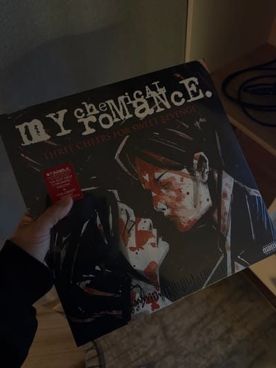 A person is holding a vinyl record album titled "My Chemical Romance: Three Cheers for Sweet Revenge". The album cover features an illustration of two figures covered in red splatters, presumably blood. A sticker on the album highlights the songs "I'm Not Okay (I Promise)", "Helena", and "The Ghost of You". The background is out of focus, showing part of a wooden shelf with black cables. There is no indication that the image was taken in Ukromne, Ukraine.