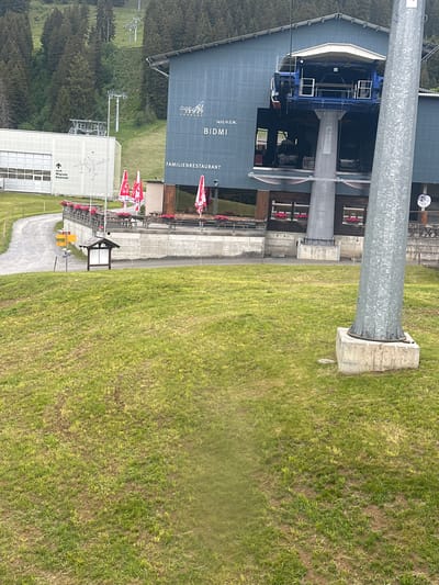 The image shows a ski resort building and a ski lift in Hasliberg Reuti, Switzerland. The building is a large, modern structure with a dark blue facade. Above the entrance, there is a sign that reads "ALPEN ROFL KRAFT", and below it, "BIDMI". The building also has a sign that says "FAMILIENRESTAURANT". A ski lift station is integrated into the building, with a large blue ski lift carriage visible. In front of the building, there is a covered outdoor seating area with tables and chairs, and red and white umbrellas. To the left of the building, there is a white structure with a garage door. A gravel path leads to this building. The scene is set against a backdrop of a green, grassy slope dotted with trees, with more ski lift structures visible further up the mountain. The overall impression is one of a functional and well-maintained tourist destination.