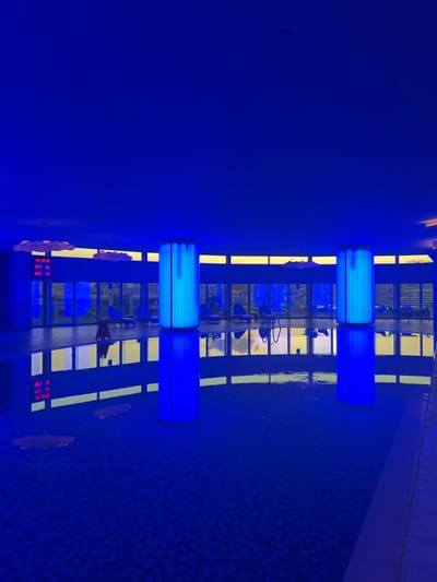 This image displays an indoor swimming pool facility in Bağcılar, Turkey, rendered in an almost monochromatic deep blue hue due to ambient lighting. The rectangular swimming pool dominates the foreground and mid-ground, its tiled bottom faintly discernible through the water. Two large, cylindrical pillars, intensely illuminated with bright blue light, stand symmetrically within the pool area, casting prominent reflections on the water's surface. Along the rear of the facility, a continuous bank of large windows or glass panels provides a view to the outside, where an obscured landscape is visible under a sky that exhibits a yellow-orange gradient, suggesting either dawn or dusk. Several blue lounge chairs or recliners are arranged on the tiled deck area beyond the pool, facing these windows. On the far left wall, above the windows, a red digital display shows numerical information: "20:38:35", "29°C 17", and "29°C 19". Multiple white, cloud-shaped decorative fixtures are mounted along the wall and ceiling line above the window array. A yellow, triangular caution sign, likely indicating a wet floor, is positioned on the pool deck to the left of the nearer illuminated pillar. No identifiable individuals are present, and no active interactions or movements are depicted. The environment is static, spacious, and appears modern.