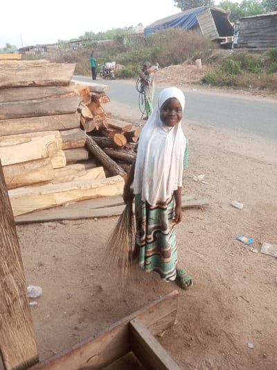 A young girl in a white headscarf and patterned dress stands on a dirt path, holding a broom. Stacks of lumber are to her left. Behind her, two figures stand near a road. One figure is on a motorcycle, while the other carries equipment. A large truck with a tarp covering the bed is visible in the background. The girl is smiling at the camera.