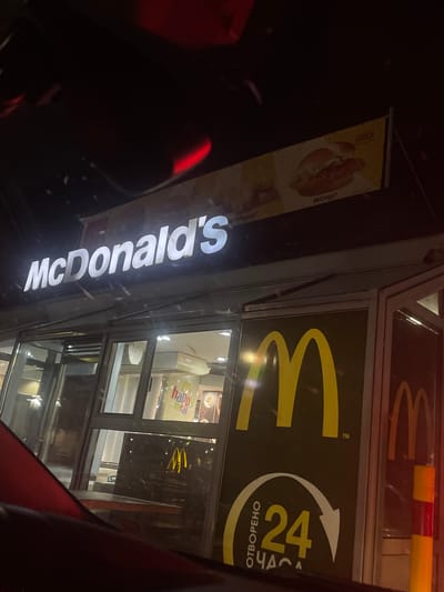 The image shows a McDonald's restaurant at night. The McDonald's sign is illuminated and prominent. The building has large glass windows and doors. Inside, there are lights and displays, including a "Happy Meal" advertisement. A large golden arch logo is visible on the exterior wall. There is also a sign indicating "24 HOURS" in Cyrillic script. The perspective suggests the photo was taken from a vehicle, with a red and black dashboard visible in the foreground. The overall scene is that of a commercial establishment in an urban or semi-urban environment.