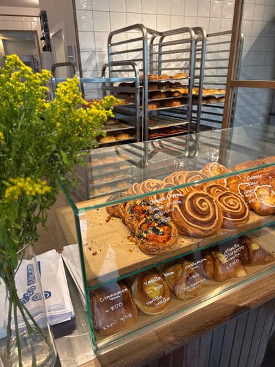 The image depicts the interior of 280 BAKARÍ, a bakery located in Reykjavik, Iceland. The scene is dominated by a clear glass display case showcasing various freshly baked pastries.
In the foreground, to the left, a tall clear glass vase holds a bouquet of green plants with numerous small, bright yellow flowers, possibly Goldenrod. To the immediate right of the vase, a stack of white paper bags with blue lettering is visible, featuring the number "280" and the word "BAKARÍ". The bags rest on a polished metal surface.
The central and right foreground is occupied by a multi-tiered glass display case with a wooden frame. The top shelf contains multiple large, swirled pastries. On the far left of this shelf, a round pastry is topped with diced red tomatoes, white feta cheese, and black seeds; a label indicates "Tomato & Feta / Tómat & Feta". Adjacent to it are several large, glazed cinnamon-roll-like pastries, with labels stating "Espresso glaze", "Espresso snúður", and one priced at "~850" (likely Icelandic Krona). The bottom shelf of the display case features more pastries. From left to right, there is a lighter swirled bun labeled "Vanilla bun / ~800 / Vanillu snúður", followed by a twisted bun labeled "cardamom bun / ~800 / Kardimommu snúður". Next to it is a croissant-like pastry with a yellow cream filling, labeled "Vanillu cornetto / Vanilla / ~850". A darker, rectangular bun is visible further left on the bottom shelf, labeled "Cinnamon bun / ~800 / Kanill snúður". The pastries are displayed on light brown paper or parchment.
In the midground, behind the display case, two tall, multi-shelf stainless steel bakery racks are visible. These racks hold numerous baking trays, many filled with golden-brown baked goods, including what appear to be croissants and other pastries, indicating a fresh production area. The wall behind the racks is covered with white square tiles. To the far left, a doorway leads into an adjacent room. No individuals are present in the frame.