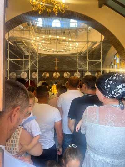 The image depicts a group of people gathered inside a building with arched entrances and brickwork, suggesting a church or similar religious structure. Scaffolding is visible overhead, indicating ongoing renovation or construction work. A large, ornate chandelier hangs prominently. Several circular icons with religious figures are mounted on the wall. In the foreground, a man is holding a young child. Other individuals, predominantly men and women, are seen from behind, facing towards the interior of the building where a person in yellow is visible. The overall atmosphere appears to be one of a religious gathering or event. The specific location in Montgeron, France, is not identifiable from the visual cues alone.