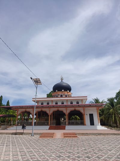 The image shows a mosque located in the city of Sawang, Indonesia. The mosque has a large black dome and is surrounded by palm trees. The building is white with orange trim and features arched walkways. In front of the mosque, there is a paved courtyard with a solar-powered street light. The sky is partly cloudy. There are no people visible in the image.