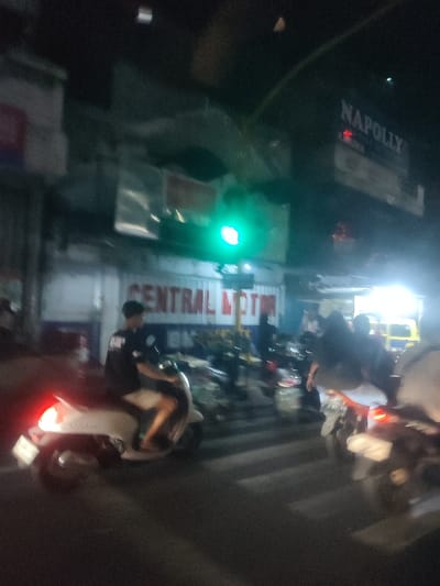 The image shows a street scene in Kecamatan Pandaan, Indonesia, taken at night. The location is identifiable due to the coordinates provided, -7.654 latitude and 112.695 longitude. There are two motorcycles on the street, both traveling in the same direction. One is a white scooter, with a man wearing a black t-shirt driving it. The second motorcycle is black and behind the white one, but it's difficult to discern the driver's identity or other details. The image shows a bright green traffic light in the distance and various other details, including a sign for "CENTRAL MOTOR". It is hard to determine the exact time, but the street is dimly lit and the time is probably around dusk or late evening. The street is paved and appears to be relatively deserted with no other vehicles visible. The image was likely taken by someone driving or walking along the street.