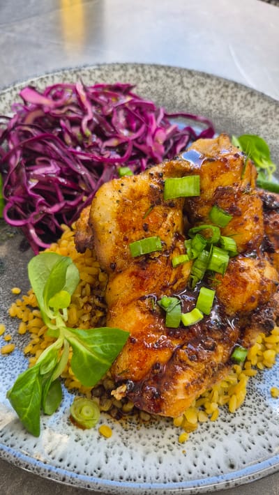 The image shows a close-up of a dish served on a grey and white speckled plate. The dish consists of several pieces of glazed chicken, topped with chopped green onions, resting on a bed of yellow grains. To the left of the chicken is a generous portion of shredded red cabbage. A few green leaves are also visible as a garnish. The lighting suggests the dish is placed on a table, likely indoors or in a covered outdoor area, with a blurred background indicating a restaurant or dining setting. The scene does not contain any information about Zarasai, Lithuania.