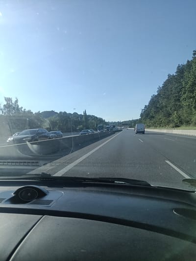 This media file captures a view from inside a vehicle traveling on a multi-lane highway on a clear, sunny day. The scene is dominated by the highway itself, with a concrete barrier separating lanes and dense green trees lining the right side of the road, suggesting a semi-rural or forested environment. The time of day appears to be late morning or early afternoon, given the bright sunlight and clear blue sky with minimal cloud cover.
Several vehicles are visible on the highway, indicating moderate traffic, with cars and trucks forming a queue in the distance on the left side and a white van in the right lane ahead. The perspective is from the driver's seat, with the dashboard and windshield wipers in the foreground, adding to the sense of being in transit.
Notable details include the sun's glare, which creates some bright spots and reflections on the windshield. The overall mood is neutral, conveying the mundane experience of highway travel. There is no visible text in the image. The location context is Kunice, Czechia, which aligns with the highway infrastructure and the surrounding vegetation depicted.