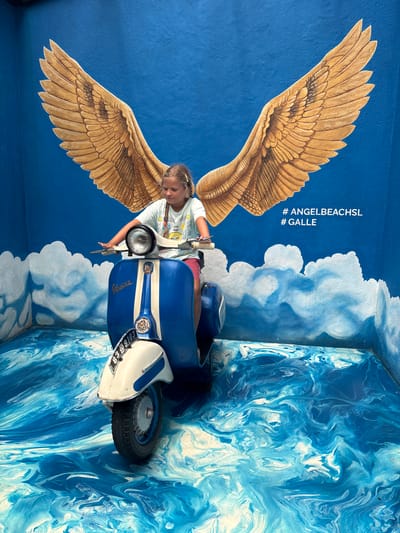 The image shows a young girl sitting on a blue and white Vespa scooter in front of a painted mural at #AngelBeachSL in Galle, Talpe, Sri Lanka. The mural consists of a blue background with painted golden wings behind where the girl sits on the scooter. The hashtag #AngelBeachSL and #Galle are also painted above the scooter. White clouds are painted below the scooter. The wall to the right of the scooter is painted in a swirl of blue and white. The scooter has a license plate that reads "83-89103." The girl is wearing a white t-shirt and red shorts. She is looking down.