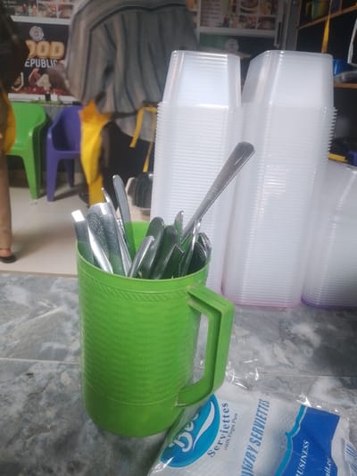 In a restaurant in Barako, Nigeria, a green plastic mug overflows with stainless steel cutlery (forks, knives, spoons) on a gray-patterned countertop. Behind the mug are stacked clear plastic containers. A sealed package of "Luxury Serviettes" is partially visible near the mug. In the background, there are patrons near chairs and a wall with a food advertisement.
