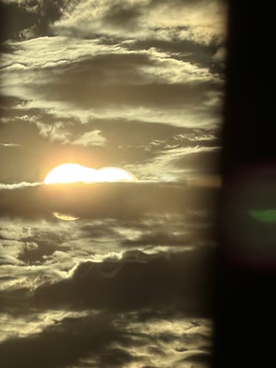 The image depicts a dramatic view of the sun partially obscured by thick, textured clouds. The sun appears as a bright, luminous orb, with a significant portion of it covered by a dark, cloud-like formation that creates a stark contrast. The clouds themselves are voluminous and wispy, with visible striations that suggest movement and depth. They are illuminated from behind by the sun, creating a radiant glow along their edges and highlighting their intricate textures.
The scene evokes a sense of awe and perhaps a touch of mystery, with the sun's light struggling to break through the dense atmospheric conditions. The overall color palette is dominated by warm tones of yellow and white from the sun and its light, contrasted with darker grays and blacks in the shadowed parts of the clouds. The composition is abstract, focusing on the interplay of light and shadow within the cloudscape. The upper portion of the image is darker, suggesting either a late hour or a deep shadow cast by an unseen element. There are no discernible people, objects, or explicit text within the frame. The overall impression is that of a powerful natural phenomenon, likely a sunrise or sunset witnessed through challenging weather conditions.