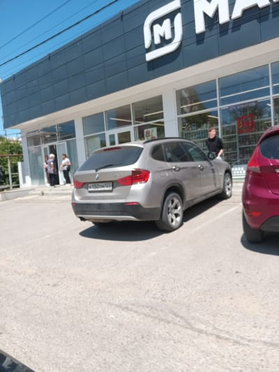 In the city of Krymsk, Russia, on a sunny day, a silver BMW X1 with license plate K150TH123 is parked in a lot in front of a modern building with a dark facade and large windows. The building has a white trim and a prominent "M" logo on its upper section. Several people are visible in front of the building, some standing near the entrance, and one man is standing by the open driver's side door of a red vehicle parked to the right of the BMW. The ground is paved with asphalt, and there is some greenery visible in the background, along with a clear blue sky.