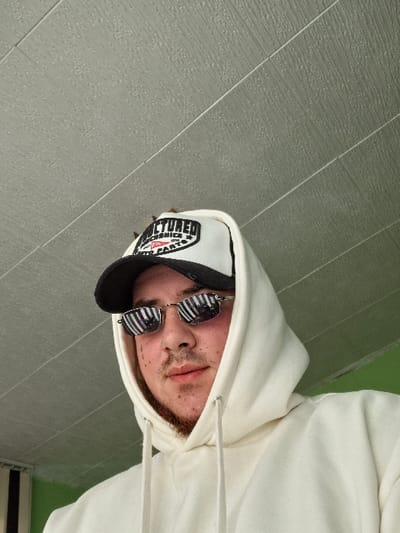 A male individual is depicted in a close-up, chest-up shot, looking directly at the camera. He wears a cream-colored hoodie with the hood pulled over his head, a black and white trucker hat with the text "STRUCTURED" and "AUTO PARTS" partially visible on the front, and reflective sunglasses featuring horizontal stripe patterns. He has a light brown beard and mustache stubble. The background consists primarily of a light grey, textured ceiling with horizontal seams. A small section of a light green wall is visible to the lower left, along with vertical black and white stripes on the far left edge of the frame. The specific location within the city of Rakitovo, Bulgaria, cannot be determined from the visual information provided in the image.