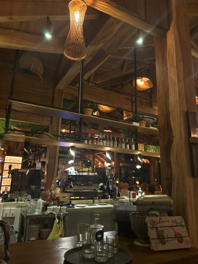 The image shows the interior of a restaurant located at latitude 9.635 and longitude -85.158 in the Cobano District of Costa Rica. The restaurant has a wooden interior with a high vaulted ceiling. There are several lights hanging from the ceiling, some of which are made of woven material. The walls are lined with shelves, which hold a variety of items, including bottles of liquor, glassware, and plants. The restaurant's kitchen is visible in the background. A countertop, which appears to be made of wood, sits in the foreground with a tray of four glasses and a sign that says "Italian Style." Behind the countertop, a stand mixer sits on the counter. There are several people visible in the background, although their faces are not clear. The restaurant appears to be relatively empty.